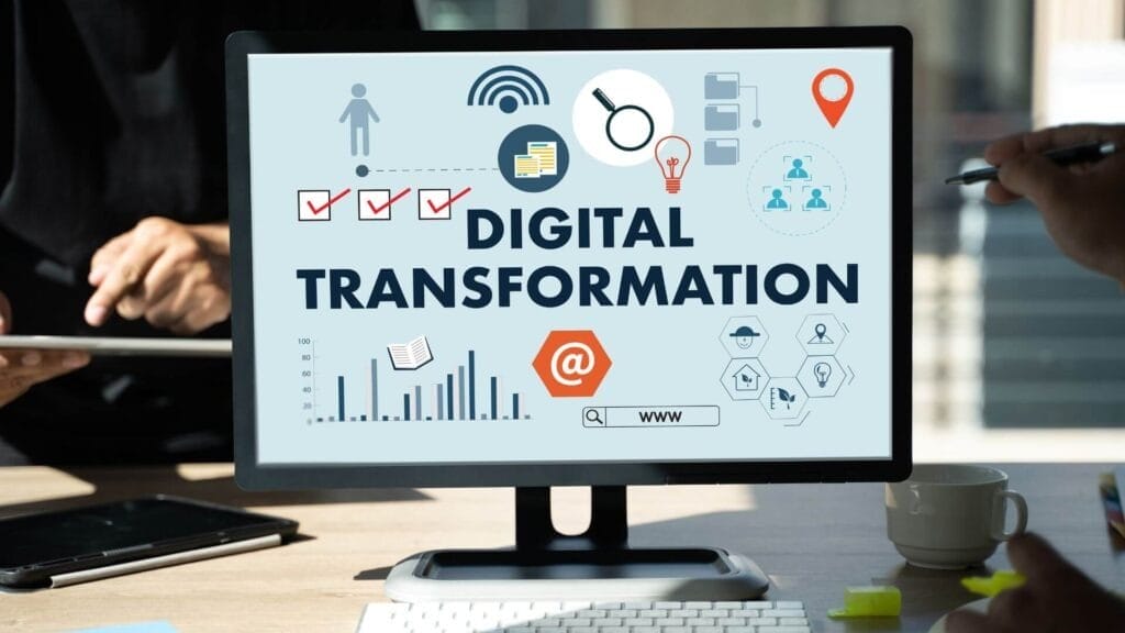 A computer screen displays the term "Digital Transformation" along with various related icons and graphics. People working around the desk are partially visible.