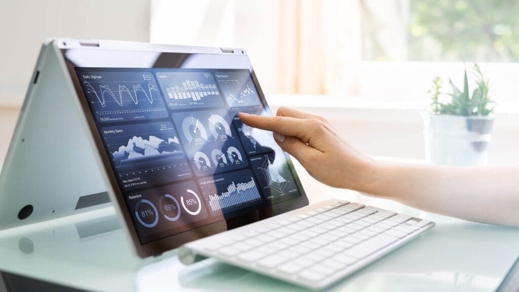 A person interacts with a touch-screen laptop displaying various data visualizations and charts. A keyboard is placed in front of the laptop on a desk.