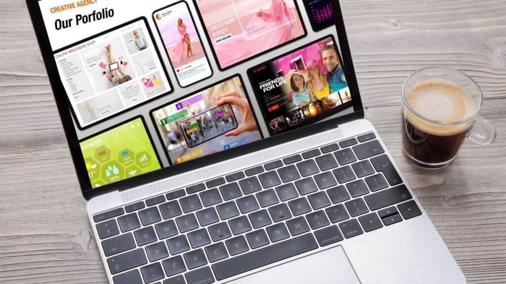 A laptop displaying various colorful digital portfolio designs on its screen sits on a wooden desk next to a clear glass cup filled with coffee.