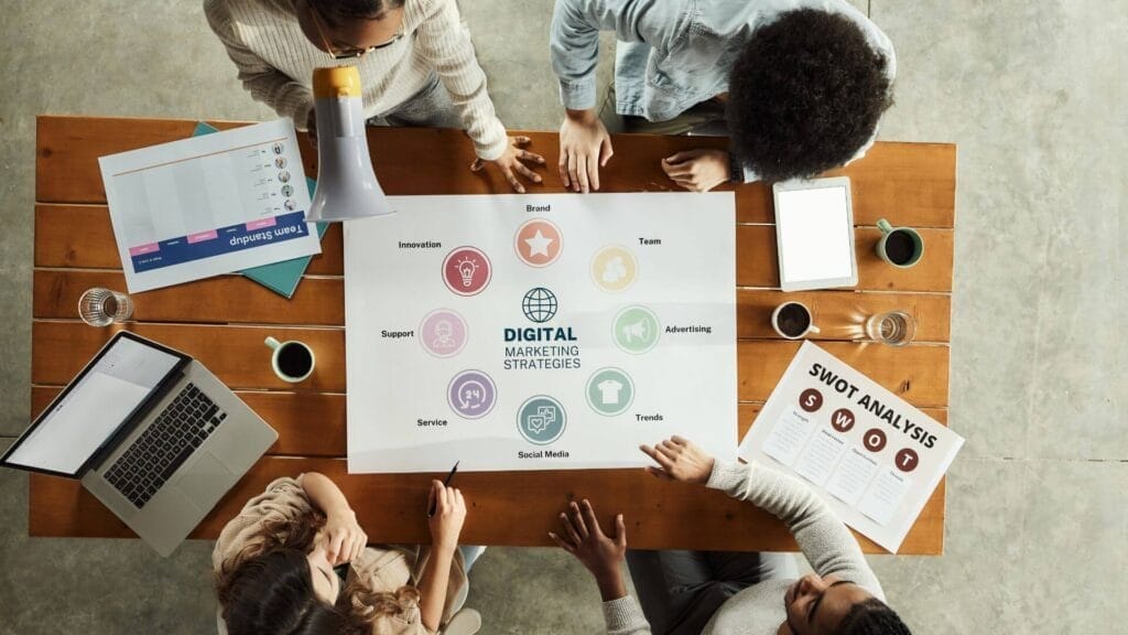 Four people sitting around a wooden table discussing digital marketing strategies using various documents and a laptop.