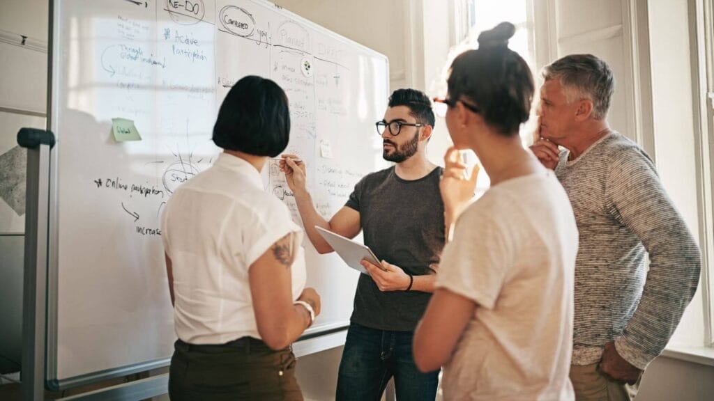 Four colleagues stand around a whiteboard, engaged in a discussion. One individual points to the board while holding a tablet. Notes and diagrams are visible on the whiteboard.